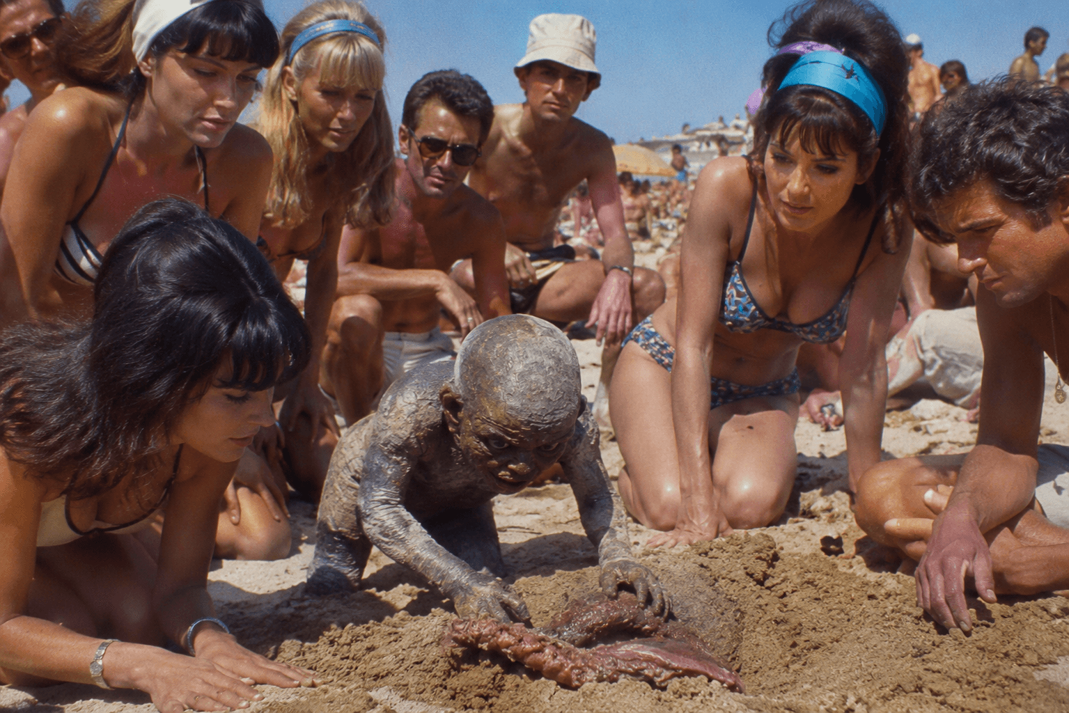 Crowd on beach gathers around mud-covered creature with tentacle.